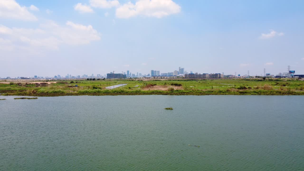 Lake and green field in foreground with Phnom Penh cityscape at distance, Drone shot