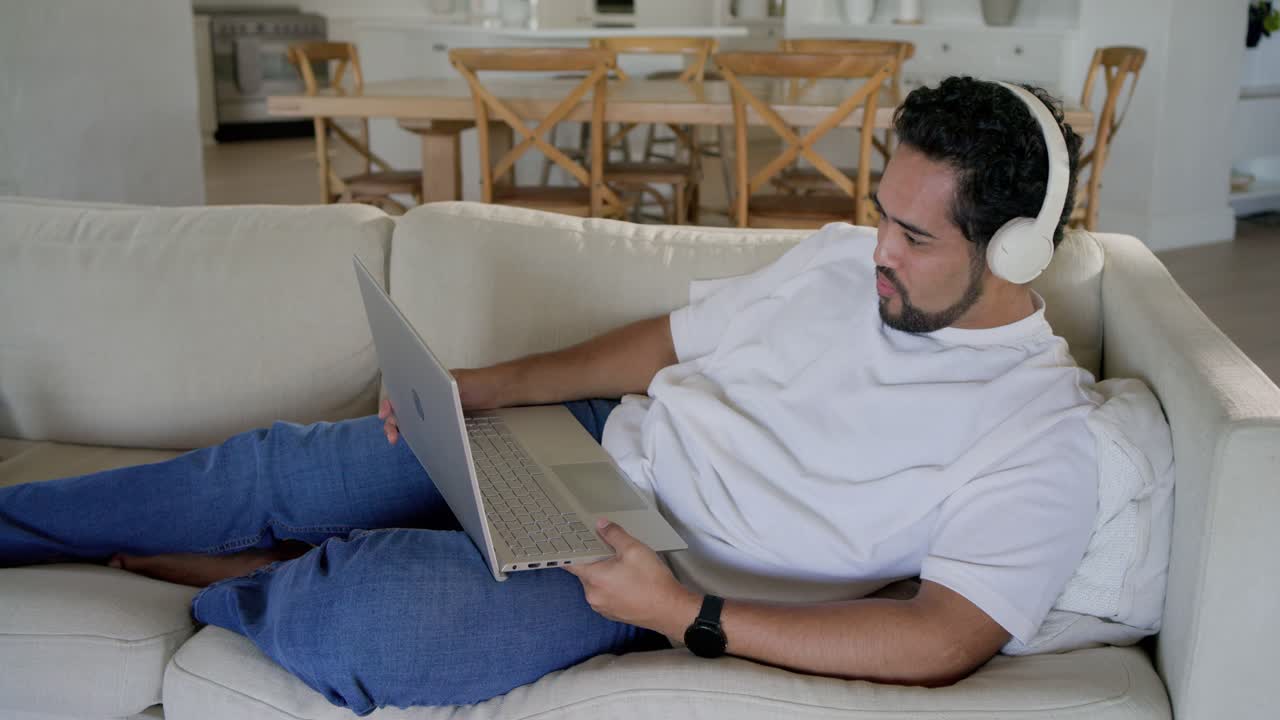 Mid-adult man lying on sofa in living room with headset, adjusting laptop while smiling at content