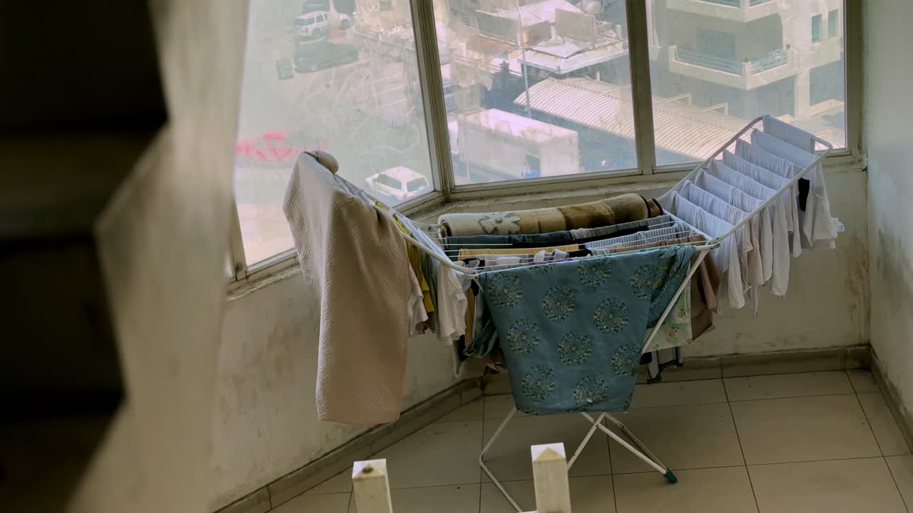 Ein Wäscheständer voller Kleidung steht neben einem großen Fenster mit Blick auf eine städtische Wohngegend, beleuchtet von sanftem Tageslicht in einem bewohnten Innenflur oder Hauswirtschaftsraum.