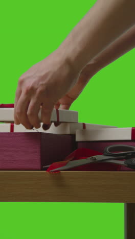 vertical video of man gift wrapping presents in boxes decorated with ribbon on table shot against green screen