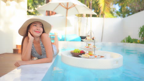 a floating buffet of deserts makes a pretty young woman lying on the edge of the swimming pool smile