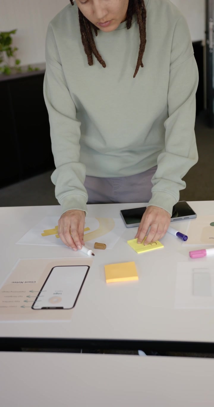 Video verticale: Persona non binaria afroamericana che scrive e allinea post-it su un tavolo accanto a un mockup.