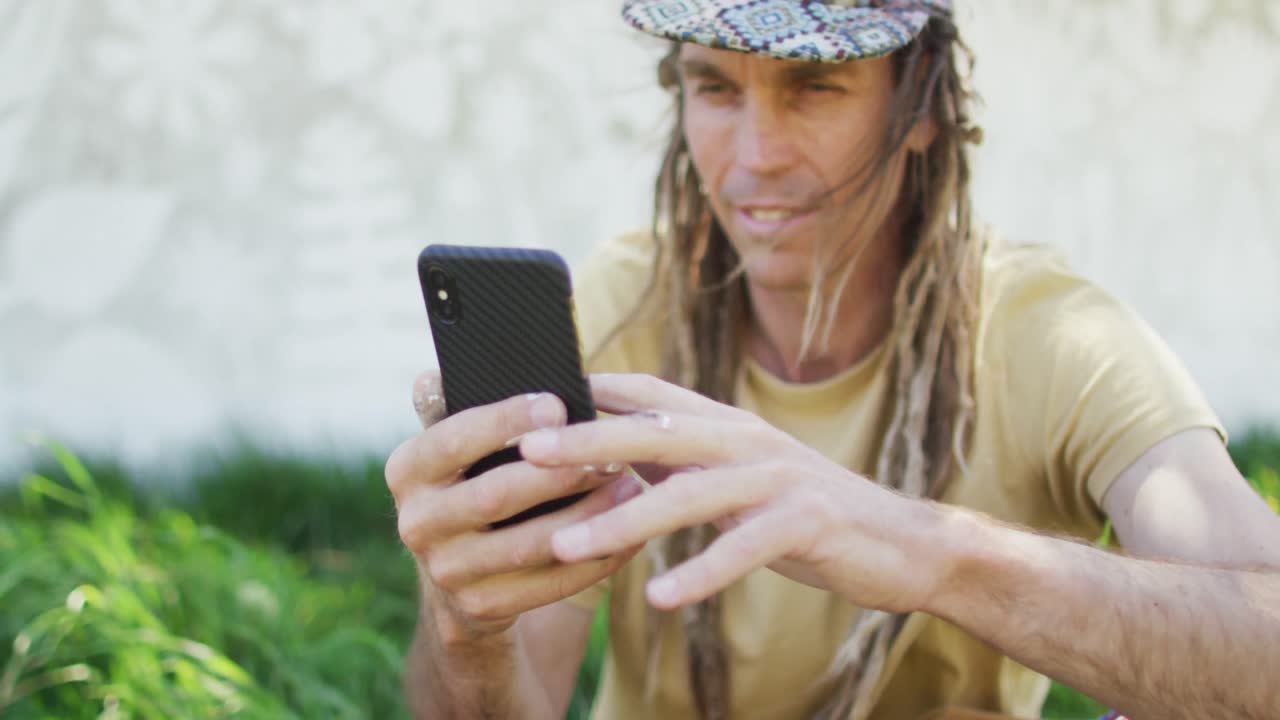 video de un hombre caucásico sonriente con rastas usando un teléfono inteligente sentado en la hierba por el mural en la pared