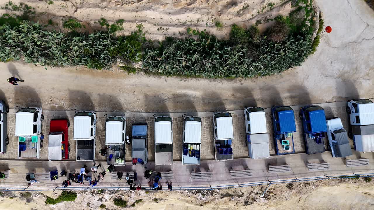 Drone Perspective of Traditional Small Trucks Parked Along a Sunny Coastal Road in Gozo Malta Showcasing the Rugged Limestone Cliffs and Vibrant Mediterranean Flora During a Bright Summer Afternoon