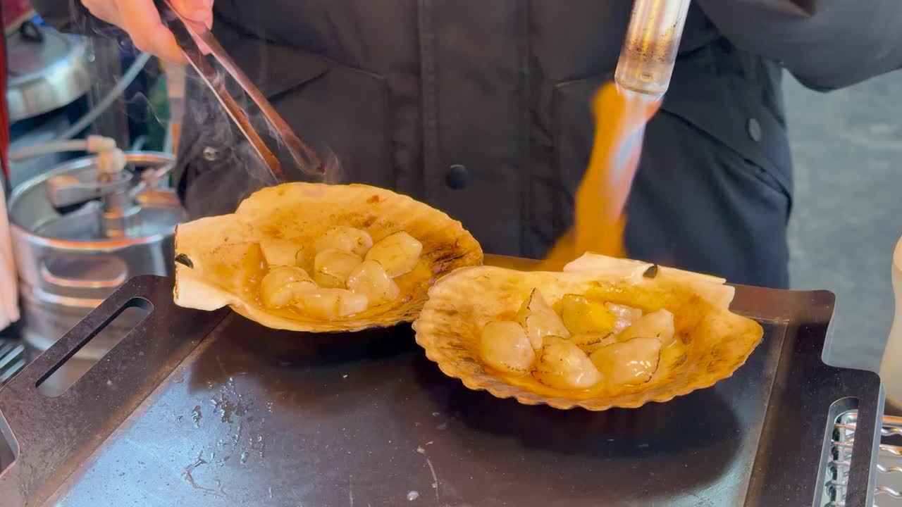 Close-up seorang penjual makanan jalanan membakar kerang simping di cangkangnya menggunakan obor las.