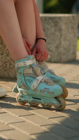 vista parcial del individuo ajustando las correas del patín de rodillos en la pierna derecha mientras está sentado, con zapatillas blancas colocadas detrás en el pavimento, vegetación en el fondo, luz solar cálida proyectando sombras suaves