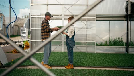 Side-view-of-a-happy-girl-Farmer-with-red-hair-giving-a-blonde-guy-with-a-beard-an-example-of-seedlings-from-a-greenhouse-during-his-work-on-the-farm