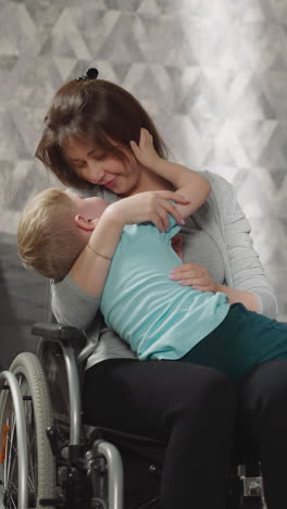 loving mother hugs and kisses toddler while daughter reading book on sofa. woman with injury sits in wheelchair expressing love to little boy in room