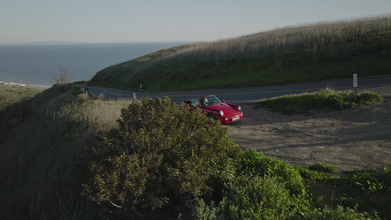 toma aérea de drones revela un porsche 1993 carrera s rojo desde detrás de un arbusto con vista al océano cerca de malibu
