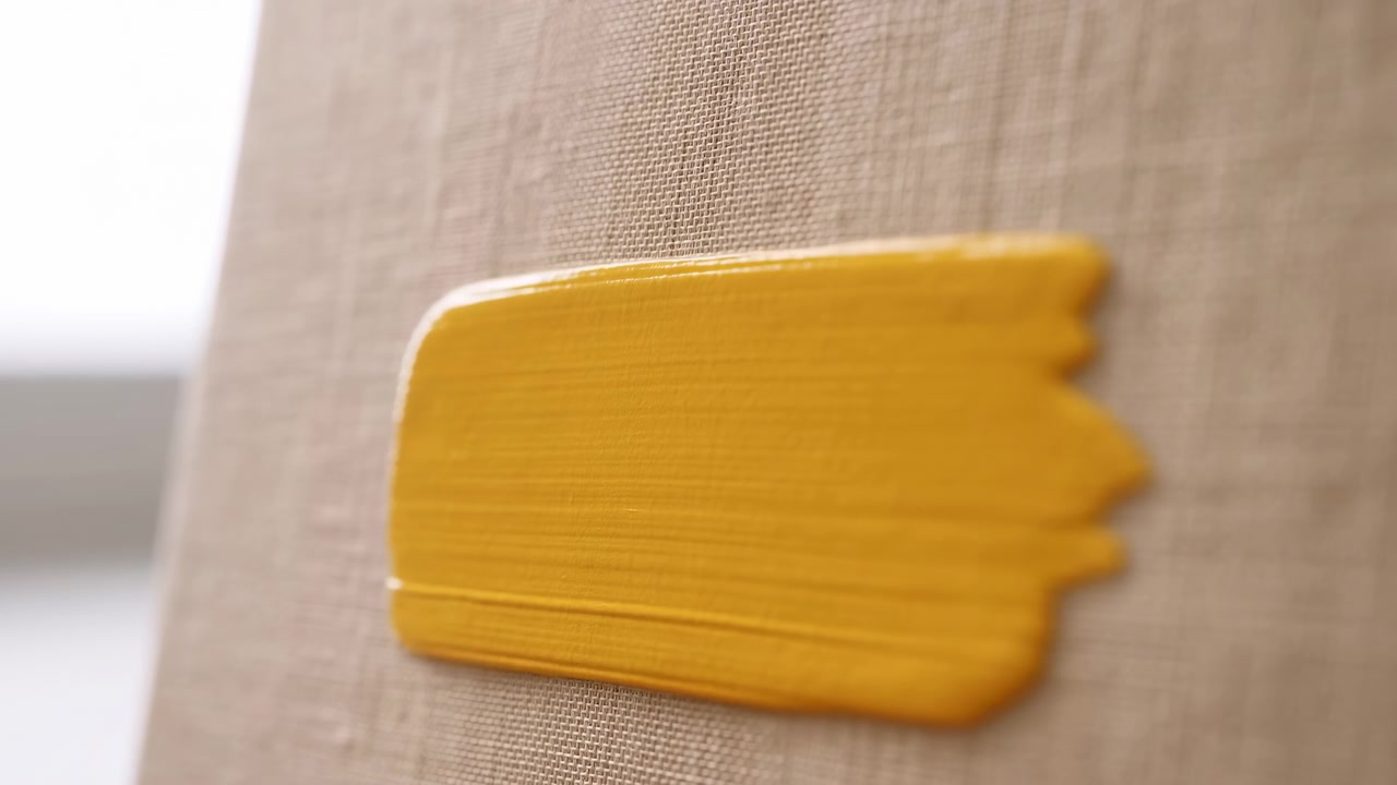 Tight oblique close-up showing freshly applied horizontal swatch of thick yellow paint on coarse beige linen canvas, with rich texture, visible weave, glossy edge, and soft studio light