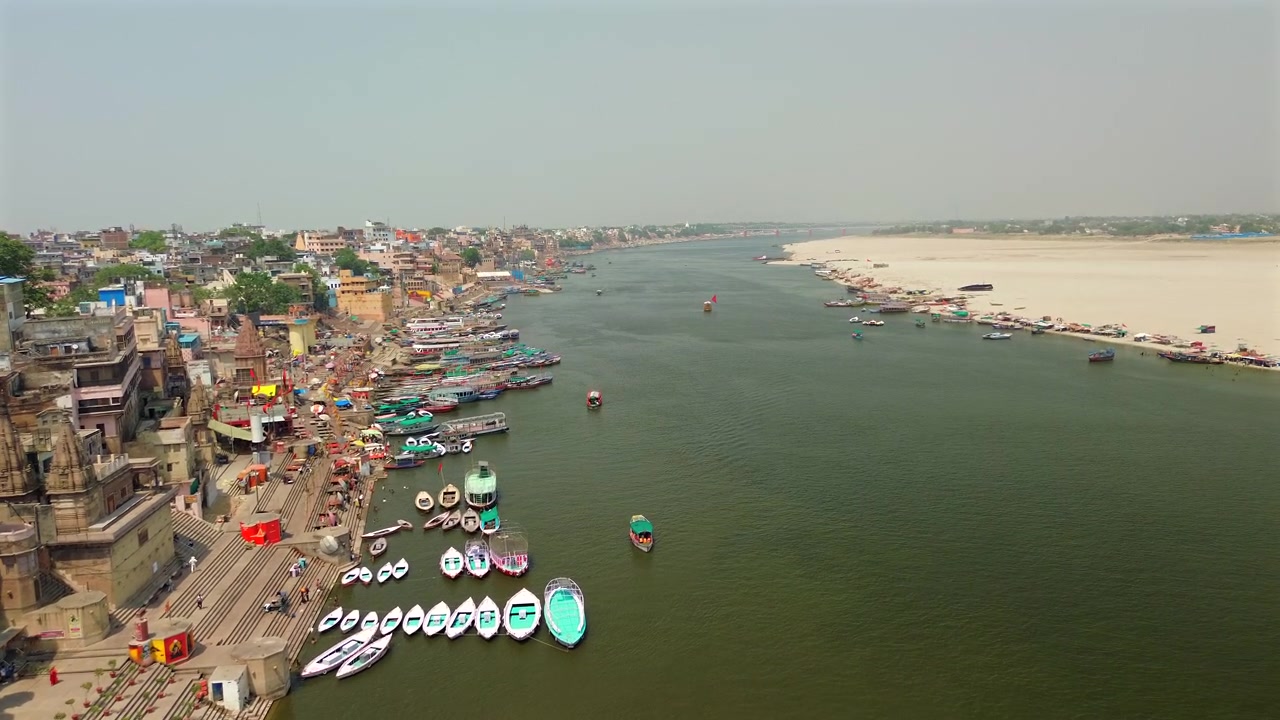 Un'ampia ripresa aerea statica che cattura il vasto e sacro fiume Gange con antichi ghat in pietra, vecchi palazzi e colorate barche di legno su un lato e una sponda sabbiosa sull'altro a Varanasi, India.