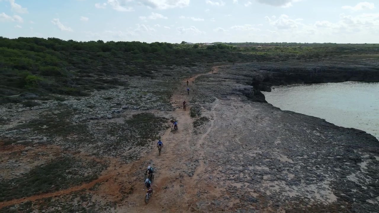 Drone Voando para Trás Revelando Litoral Épico com Ciclistas em Corrida por Trilhas de Cascalho