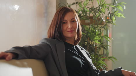 confident businesswoman sitting comfortably on couch in modern office, wearing professional attire and relaxed posture. smiling with calm demeanor, surrounded by plants in a bright office space
