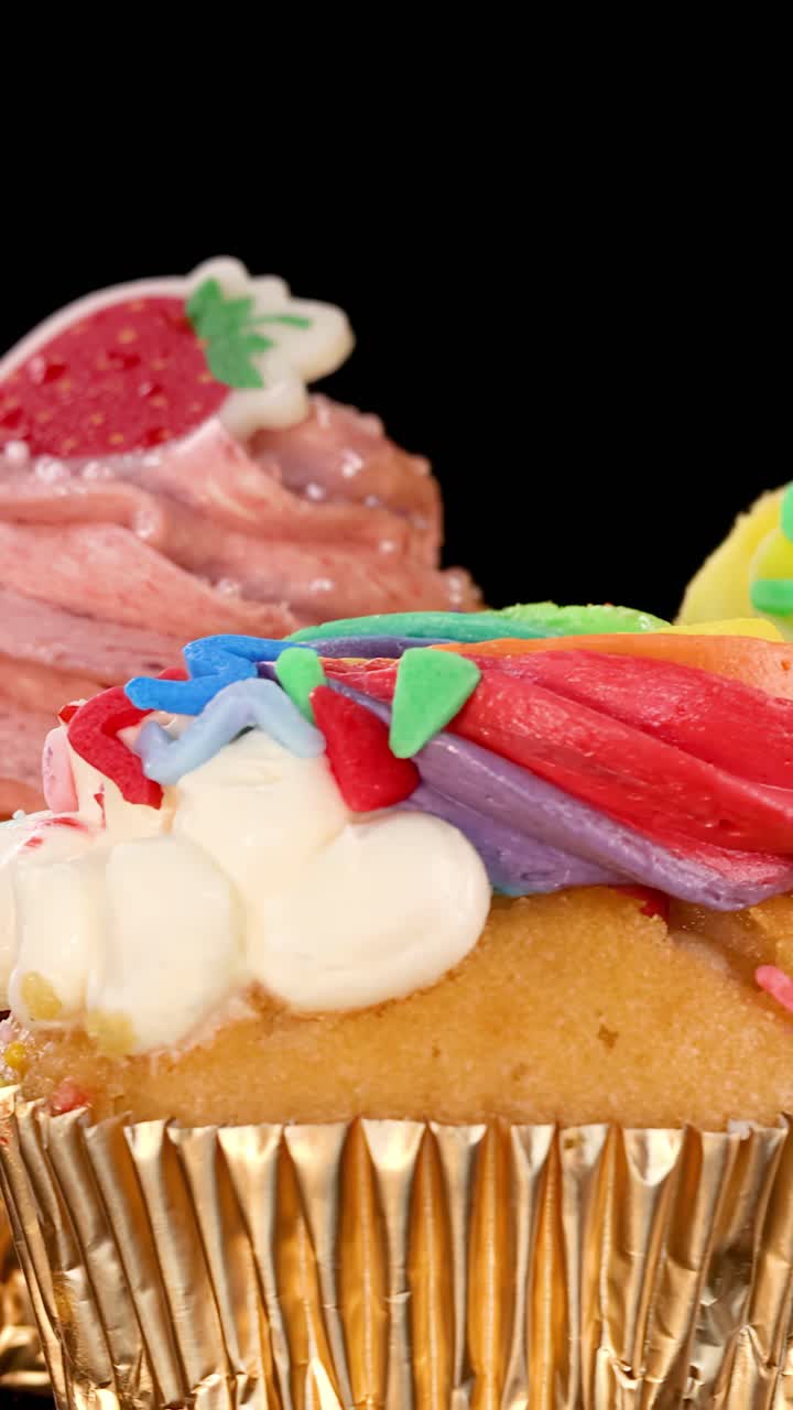 Tre cupcake riccamente decorati ruotano su uno sfondo nero, mettendo in mostra glassa vivace, zuccherini e pirottini dorati sotto un'illuminazione da studio brillante e uniforme.