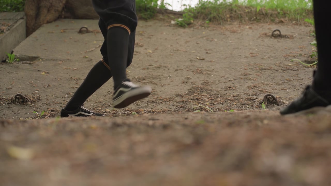 Primo piano delle gambe e dei piedi di una persona in abiti neri che cammina su terreno sterrato con foglie sparse, anelli di metallo incastonati nel sentiero e vegetazione visibile sullo sfondo. La calzatura è coerente in tutta