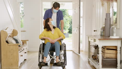 Mature-Asian-man-pushing-wife-in-wheelchair-in-hallway-at-home-both-wearing-face-masks-during-health-pandemic---shot-in-slow-motion