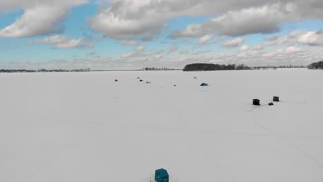 Pescador-Pescando-En-El-Hielo-Con-Cabañas-En-La-Bahía-Congelada-De-Sodus-Del-Lago-Ontario-En-El-Condado-De-Wayne,-Nueva-York,-Estados-Unidos