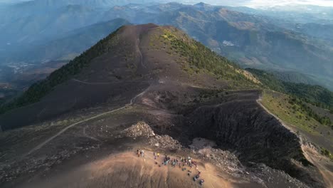 Drone-view-in-Guatemala-flying-back-over-a-volcano-crater-at-sunrise-surrounded-by-green-mountains-covered-in-ashes