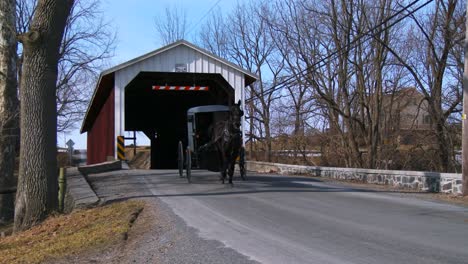 an amish horse cart travels through a covered bridge along a road in rural pennsylvania