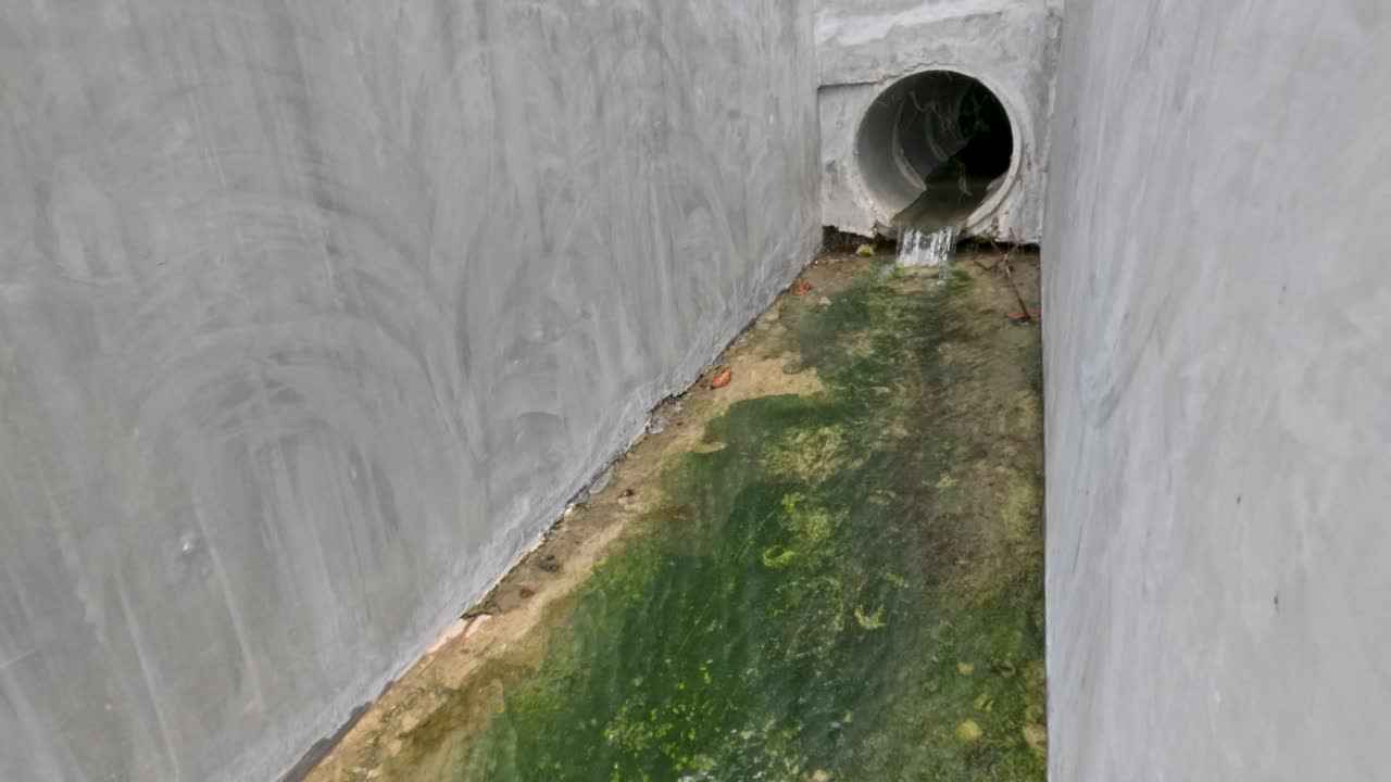 Contaminated water with plastic waste flows from concrete tunnel to rocky beach, overcast daylight