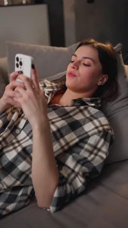 Vertical-video-of-an-enraged-and-irritated-brunette-girl-in-a-checkered-shirt-lies-on-a-gray-sofa-and-communicates-using-her-new-smartphone-in-a-modern-apartment