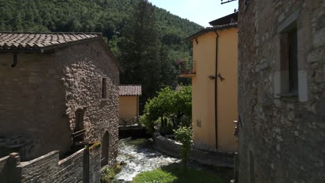 Aerial-Flying-Over-Water-Flowing-Past-Under-Wooden-Bridge-In-Rasiglia,-A-Small-Village-Located-In-The-Province-Of-Perugia