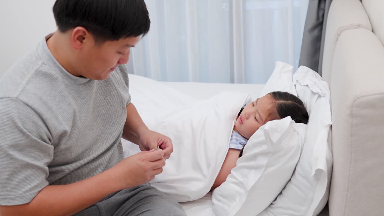 Father checks temperature and comforts sick daughter in softly lit, cozy bedroom setting