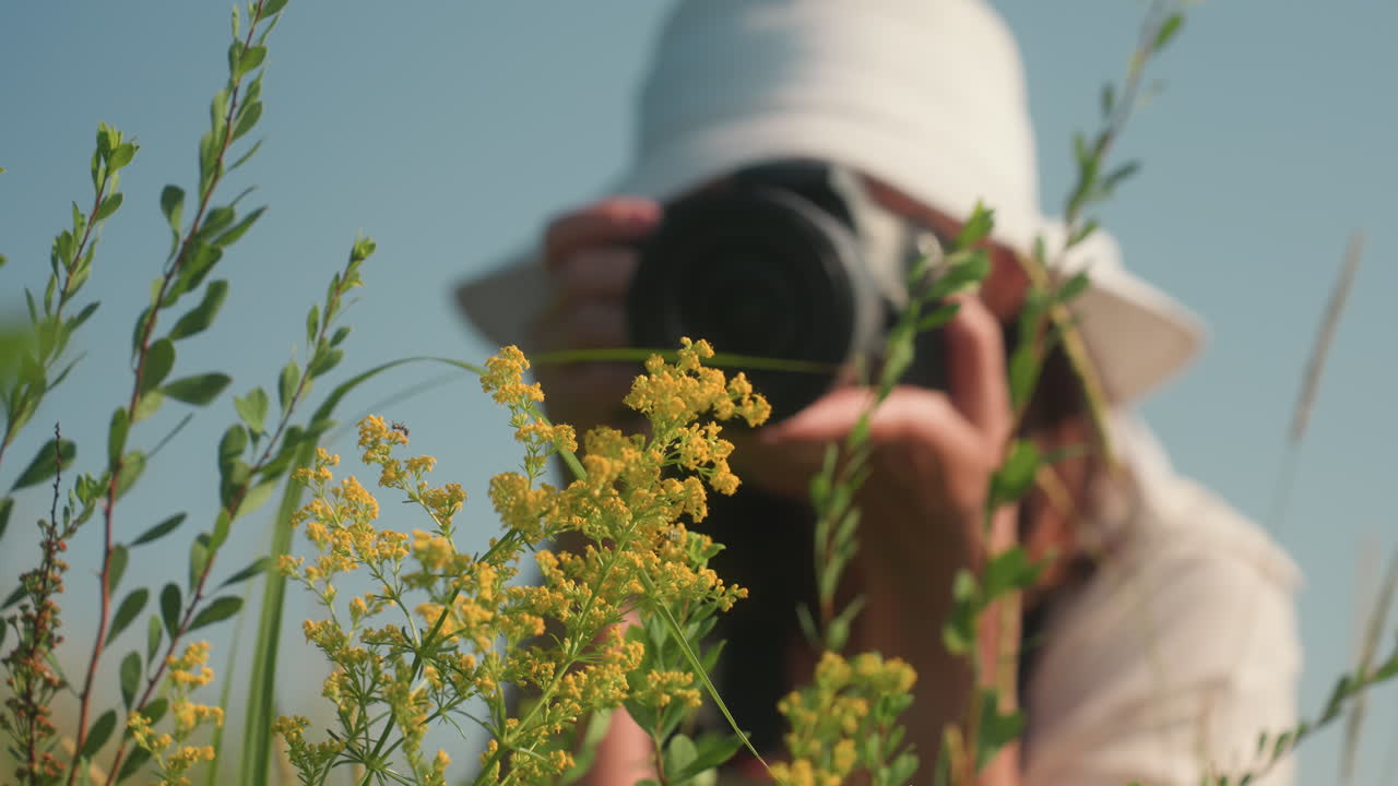 Fotógrafo com chapéu de sol branco agacha-se para capturar flores silvestres amarelas vibrantes com a câmera, mãos firmes, rosto borrado ao fundo, vegetação iluminada pelo sol emoldurando o tema floral contra um céu azul suave