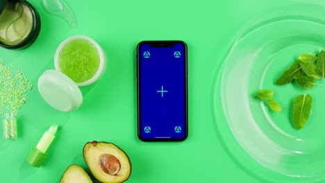 phone with green screen chroma key with fresh avocado, green cream, lipstick and essential oil on green background close-up, top view with mint mentol, make up cosmetics