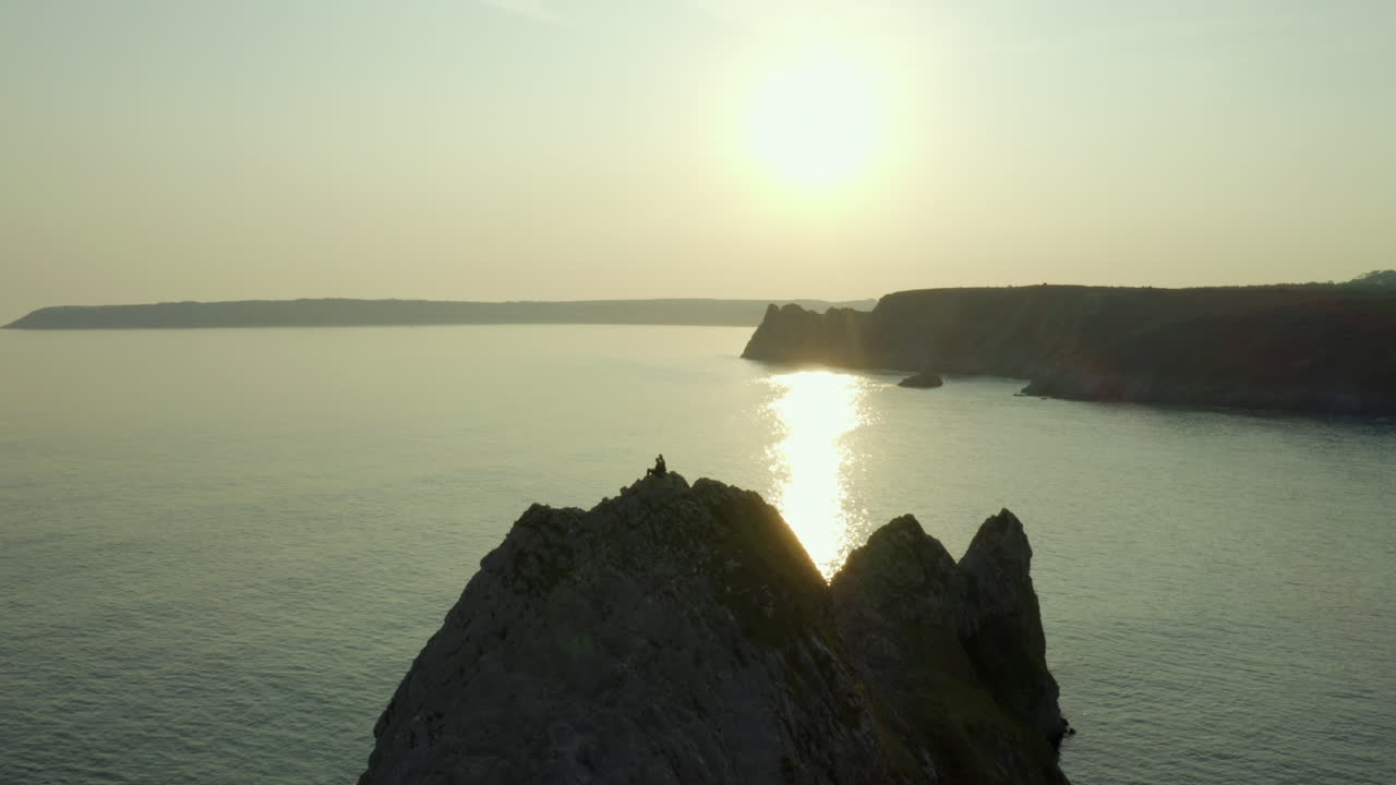 drone volando más allá de una pareja en los acantilados rodeados por el mar en la bahía de los tres acantilados con la puesta de sol en el fondo en gower, gales del sur