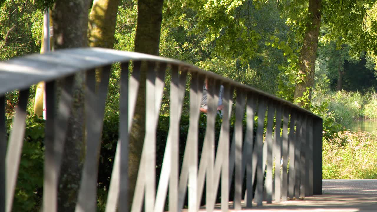 A person rides a bicycle across a sunlit bridge surrounded by lush green trees