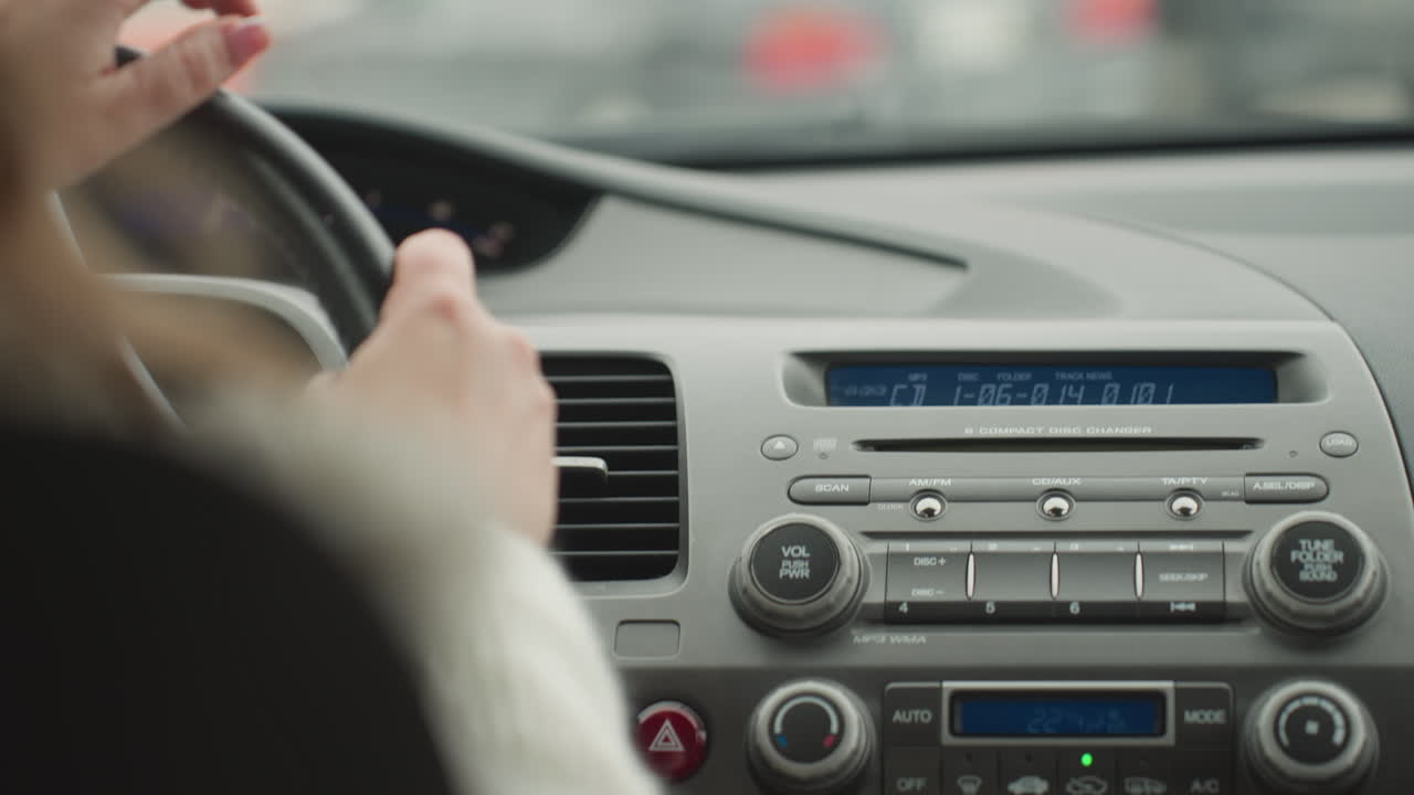 Close up of person holding steering wheel while driving during road trip with dashboard controls visible inside car and blurred view of snow-covered parked car ahead outside windshield