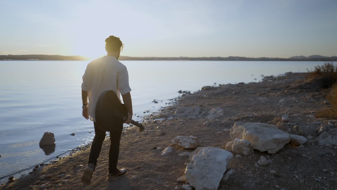 Pria Berjalan dengan Gitar di Pantai Danau saat Senja