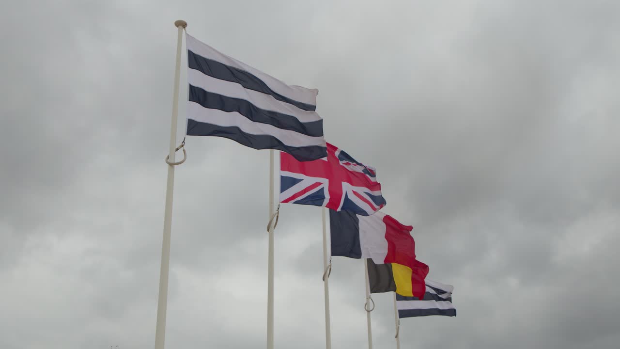 Multiple national flags wave vigorously on tall poles under cloudy skies at coastal landmark