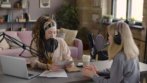 two women recording a podcast talking into a microphone sitting at desk with laptop and documents 1