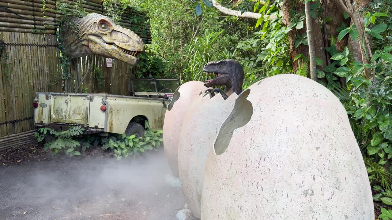 Animatronic dinosaur hatches from egg, surrounded by mist, lush plants, and old vehicle