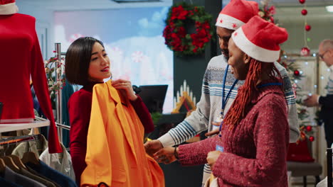 asian retail assistant and coworker helping indecisive customer choose between two colors of blazers in christmas decorated clothing shop. employees assisting client during winter holiday season