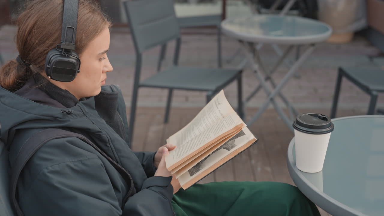 Белая женщина читает книгу на улице, в наушниках и пуховике, сидит за столиком на террасе кафе на деревянном настиле, потягивает кофе из стакана на вынос, медленно перелистывает страницы, спокойная и задумчивая