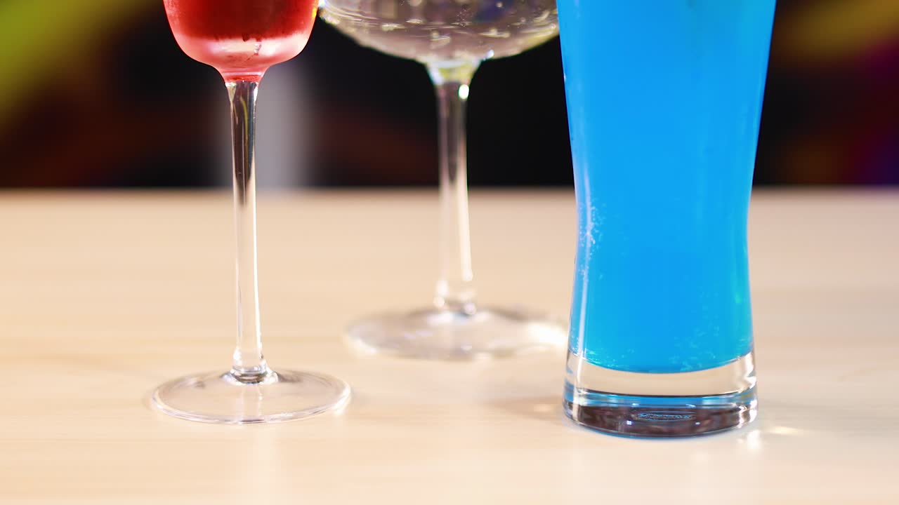 Three cocktails in vibrant colors on a table with a blurred, colorful background. Bright lighting highlights the drinks' vivid hues