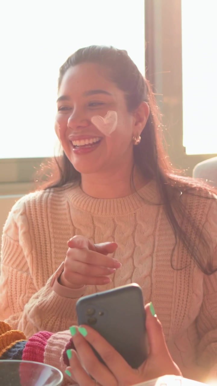 Video verticale: Donna sorridente con maglione color cammello che applaude, guardando un'amica mostrare il telefono sul divano con un barattolo.