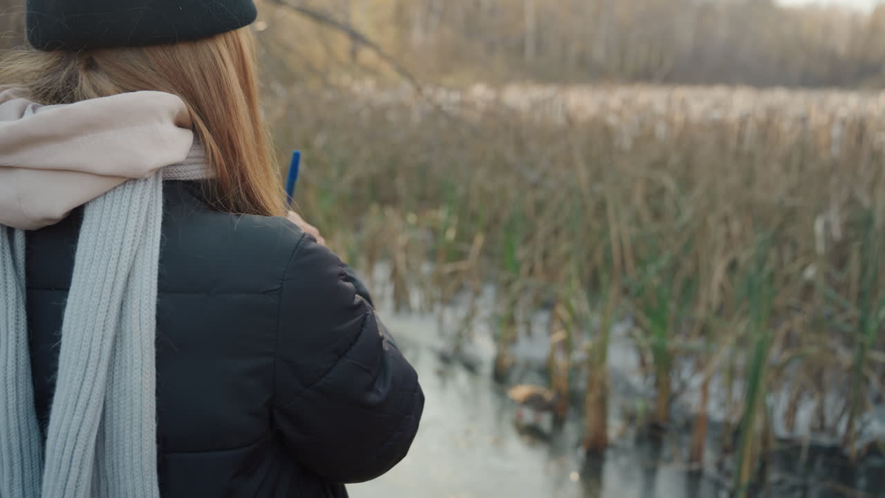 White woman taking field notes at marsh during winter light, wearing black coat, knit hat and scarf, using tablet and pen to record observations, focused researcher posture, reeds and shallow water