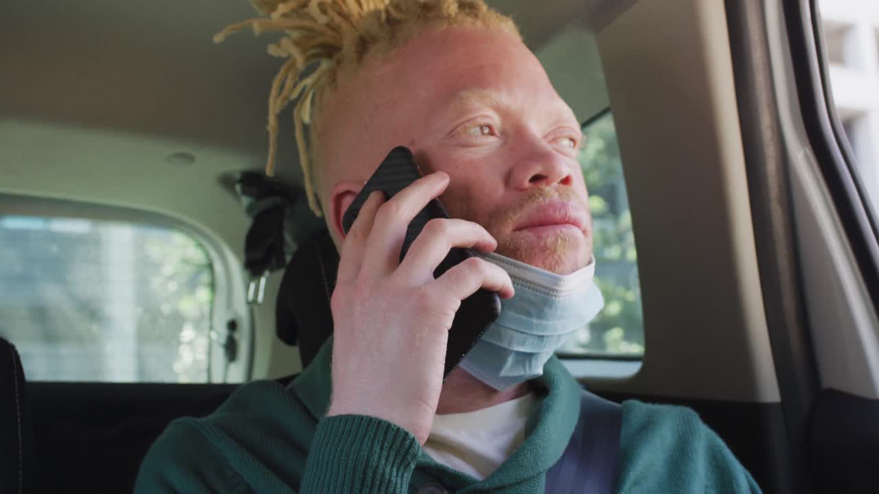 Afroamerikanischer Mann mit Albinismus trägt eine Gesichtsmaske, sitzt im Auto und telefoniert mit dem Smartphone.