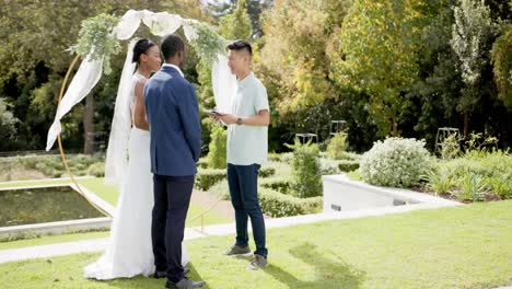 Hombre-Birracial-Oficiando-Ceremonia-De-Boda-Para-Una-Pareja-Afroamericana-En-El-Jardín,-En-Cámara-Lenta