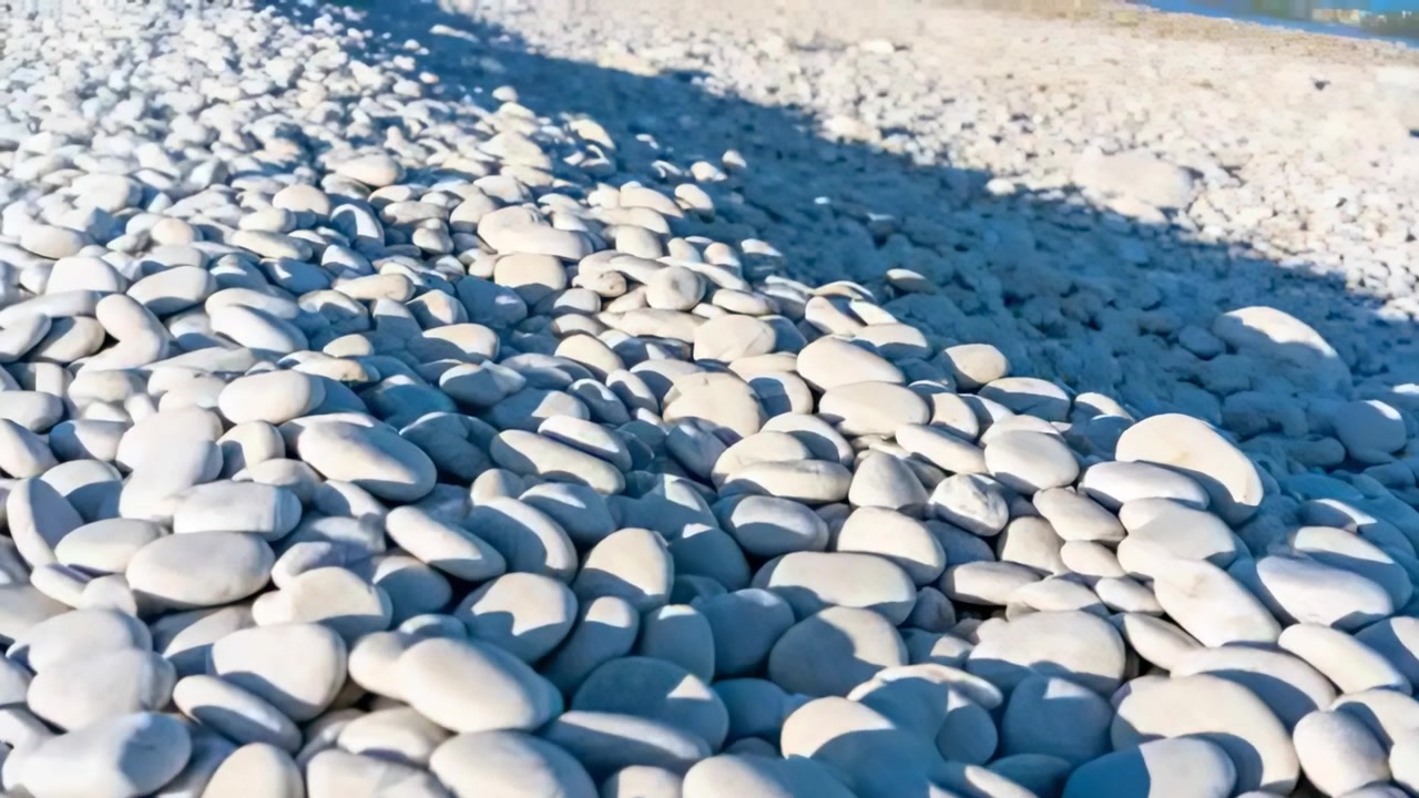 Eine schwenkende Kamera bewegt sich langsam über einen Kiesstrand und zeigt Schatten sowie den Meereshorizont.