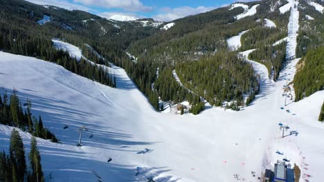 drone flies above a vibrant big sky ski resort in peak winter, capturing skiers on the lift and others racing down fresh trails, framed by pine trees and towering snow-covered mountains