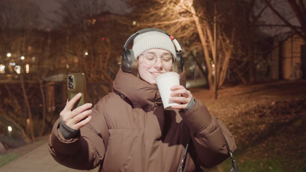 Influencer asiatica in un selfie notturno al parco con caffè, tiene telefono e tazza mentre gesticola, indossa un cappotto oversize, cuffie e berretto. Sorso giocoso e sorriso, con il bagliore del lampione e alberi spogli, scatto spontaneo.