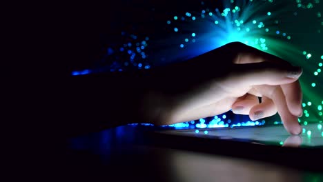 woman hand touching and browsing on tablet device in dark room with colorful blurry fiber optic lights closeup panning motion