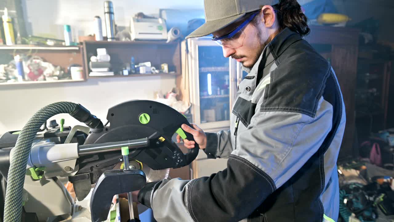 un joven con barba en un mono gris de profesión un carpintero trabaja con una máquina de corte circular en su taller de casa