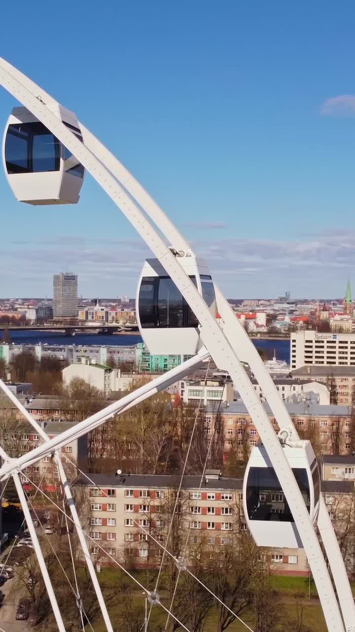 Smooth vertical view of Ferris wheel spinning with scenic Riga backdrop.