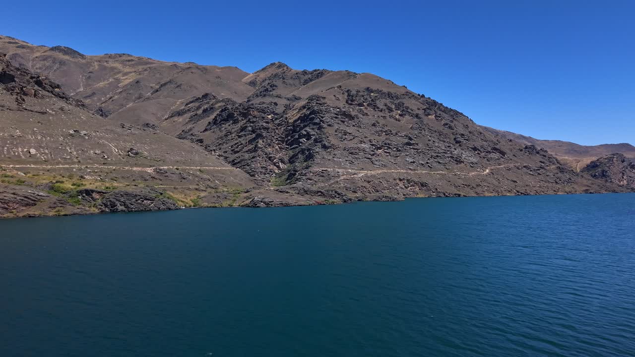 On a bright sunny day, the drone flies over the Clutha River and Dunstan Trail, capturing the winding path along rugged hills in Central Otago, New Zealand
